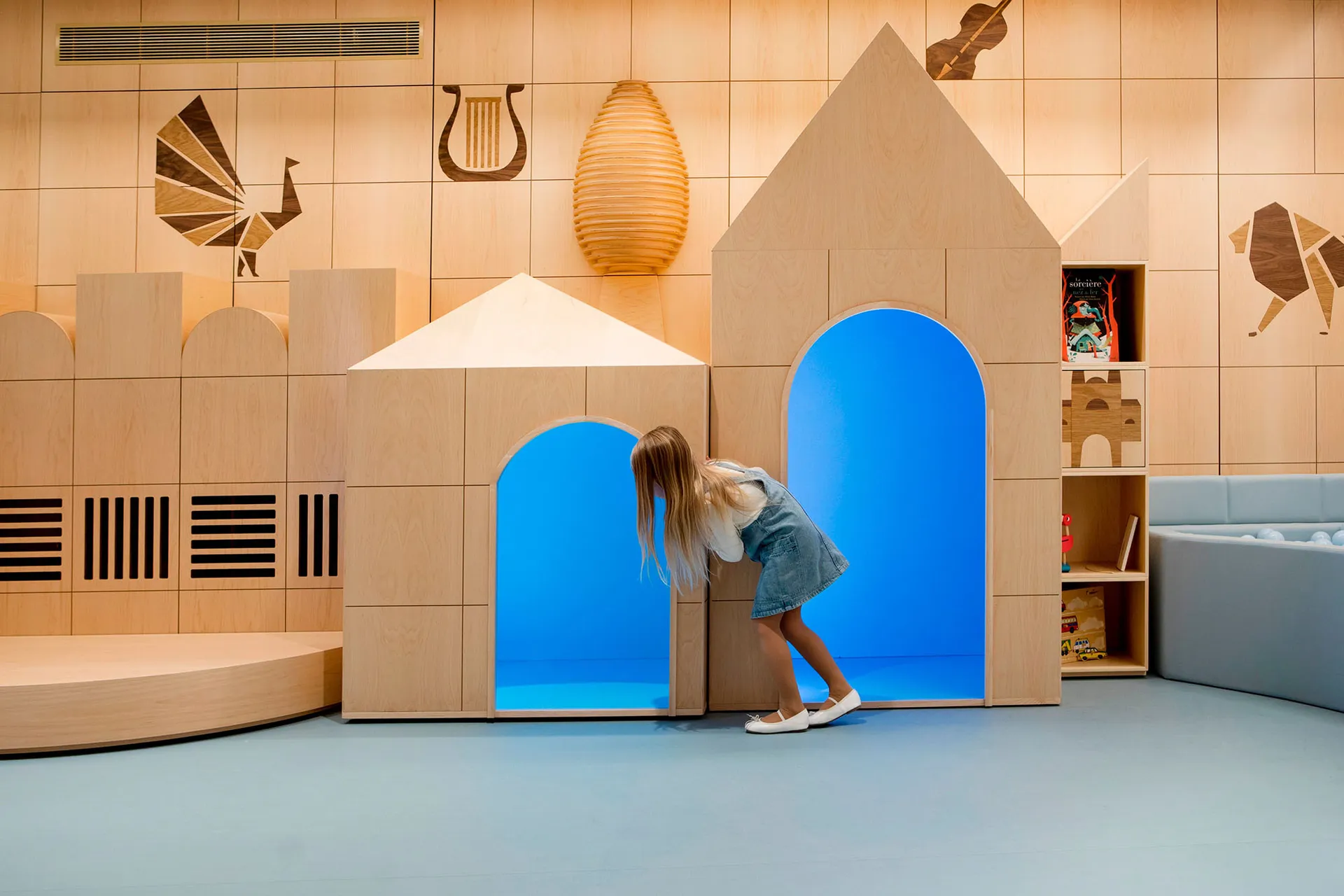 A child plays in an indoor play space featuring wooden playhouses with blue interiors.