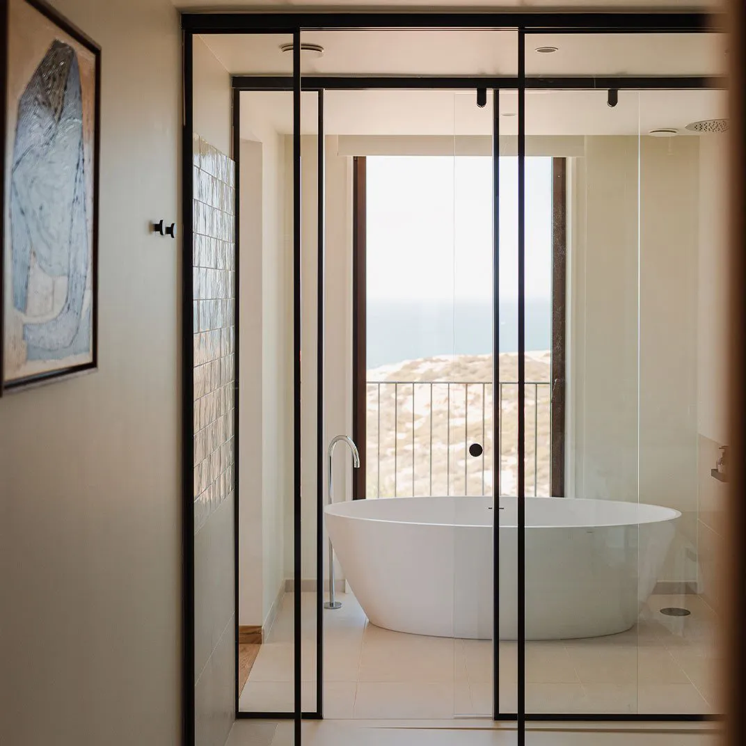Minimalist bathroom featuring a white tiled floor and walls, with a large oval-shaped bathtub overlooking the ocean.