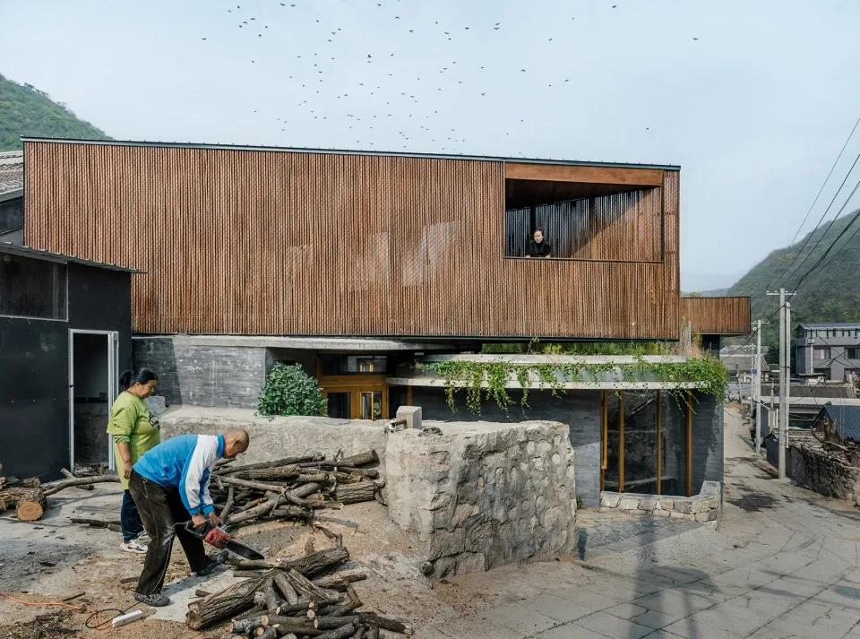 People cutting logs with a chainsaw in front of a modern building with wooden exterior and concrete walls.