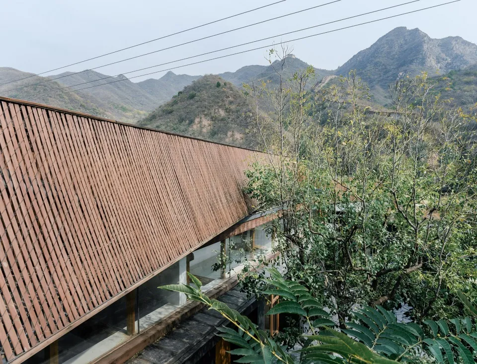 A modern architectural exterior featuring a wooden slat roof and glass walls surrounded by greenery and mountains in the back