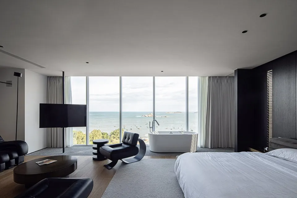 A modern bedroom featuring concrete walls and a large window with a view of the ocean.