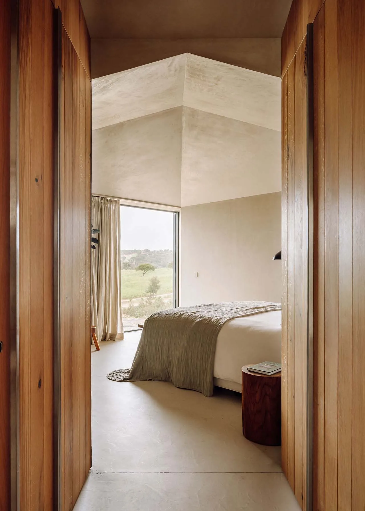 A minimalist bedroom featuring a bed with a wooden frame and a concrete floor, with a view of a natural landscape through a l