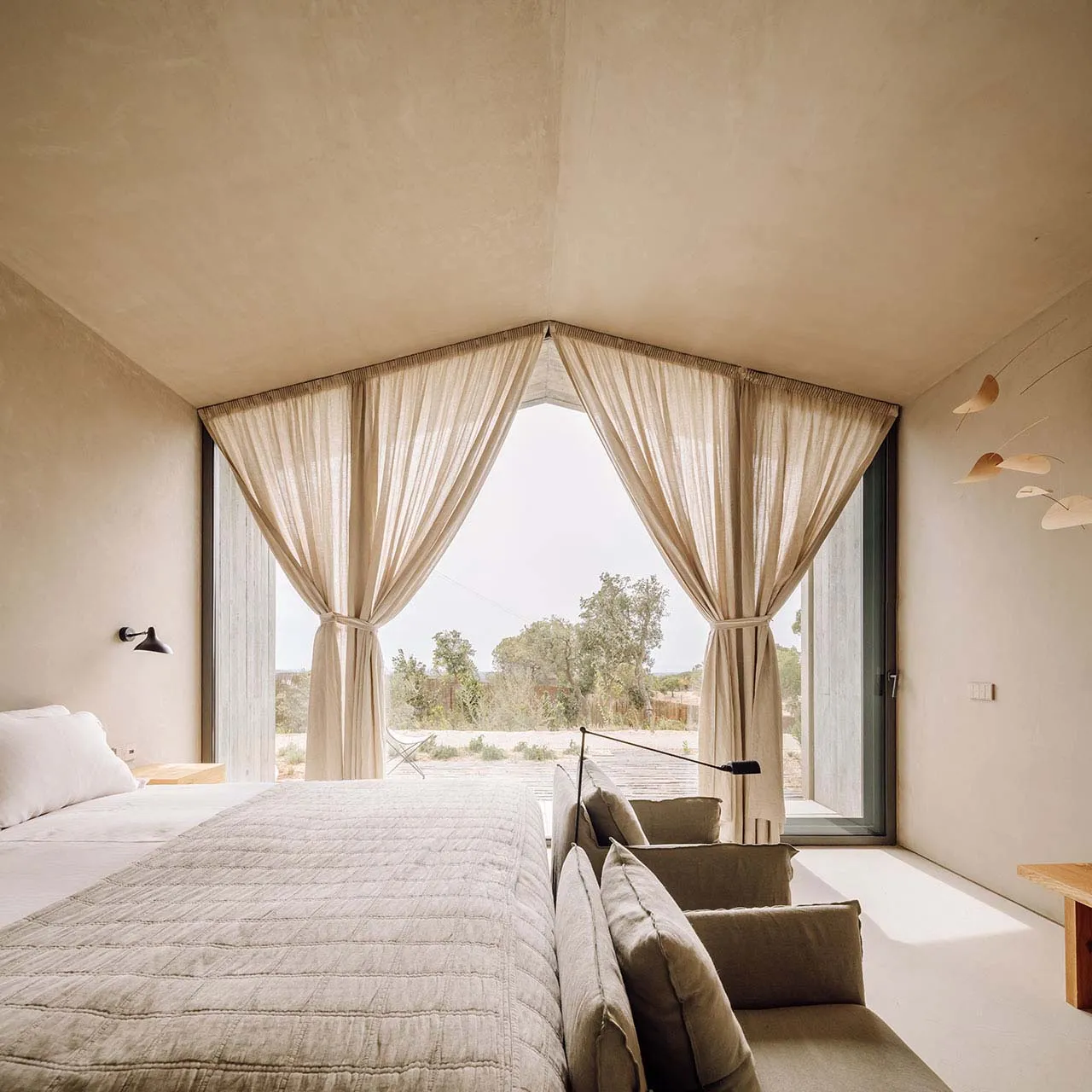 A minimalist bedroom with fabric curtains and a concrete floor, featuring a comfortable bed and a small seating area.
