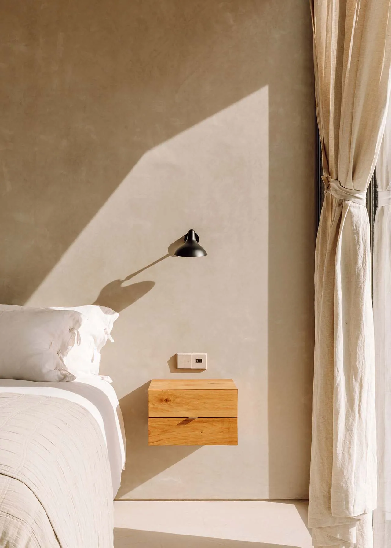 Minimalist bedroom with a wooden floating nightstand, window with curtains, and soft lighting from a wall-mounted sconce.