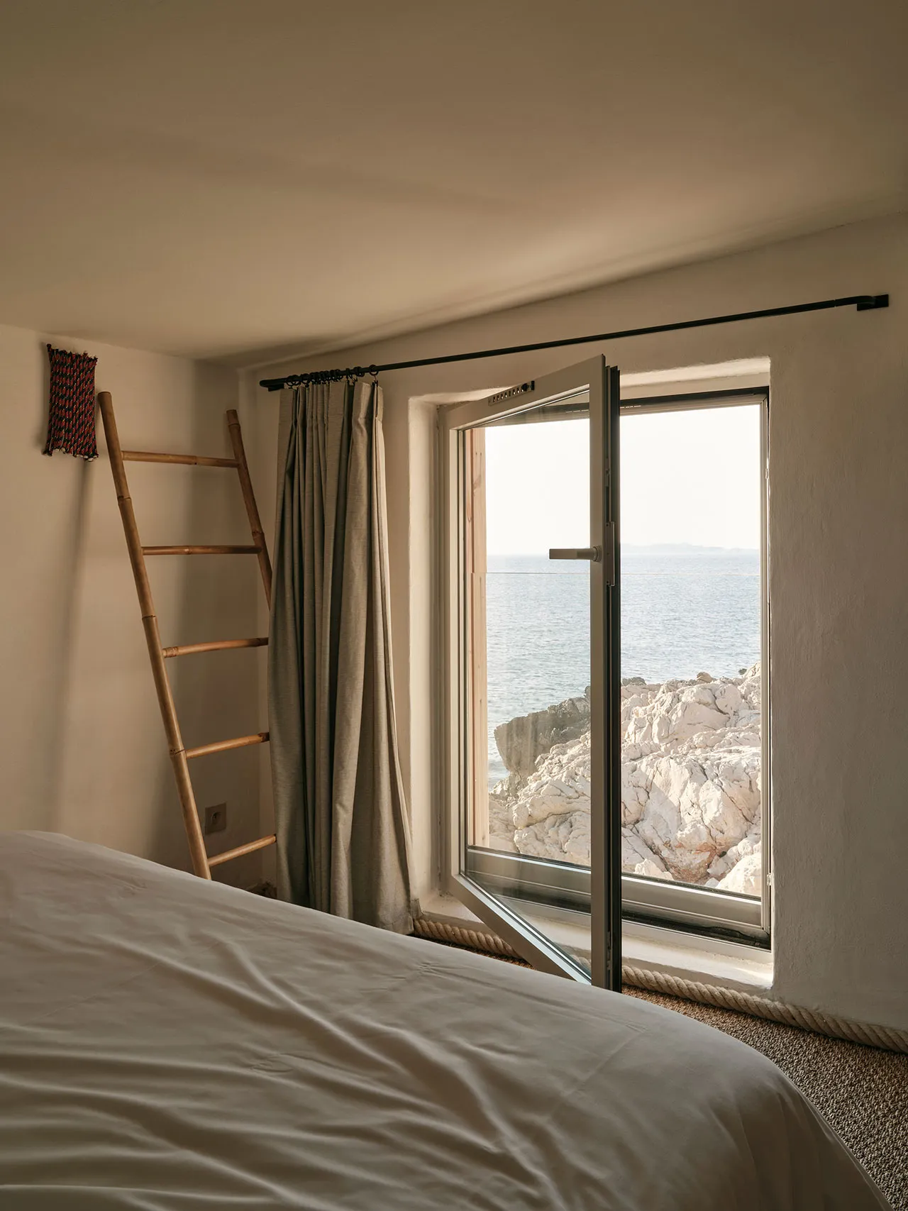 A minimalist bedroom featuring a stone construction, with a window overlooking the sea and rocks, and a bed with a white shee