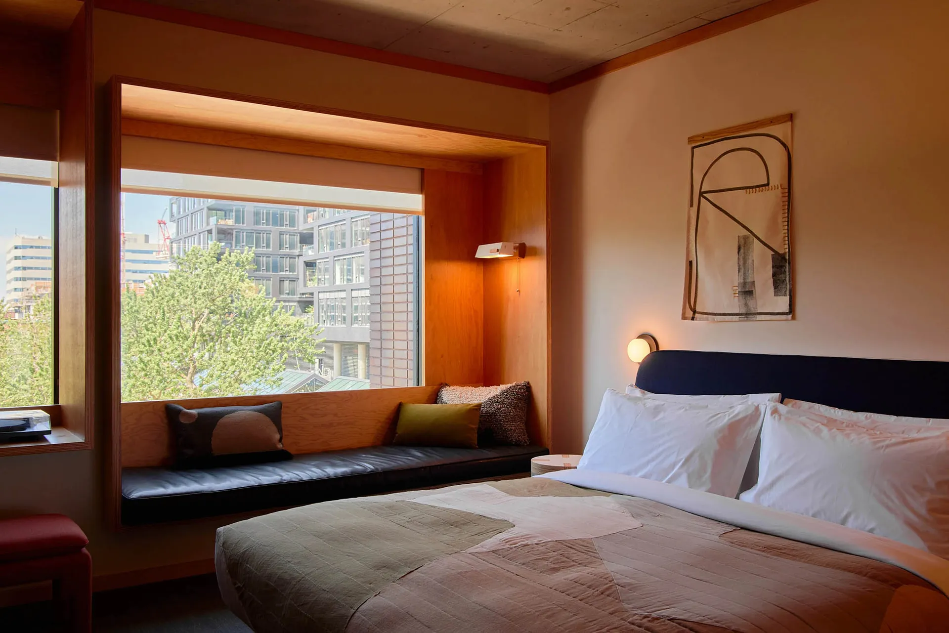 A minimalist bedroom featuring a bed with white linens, wood furniture, and a window seat with pillows.