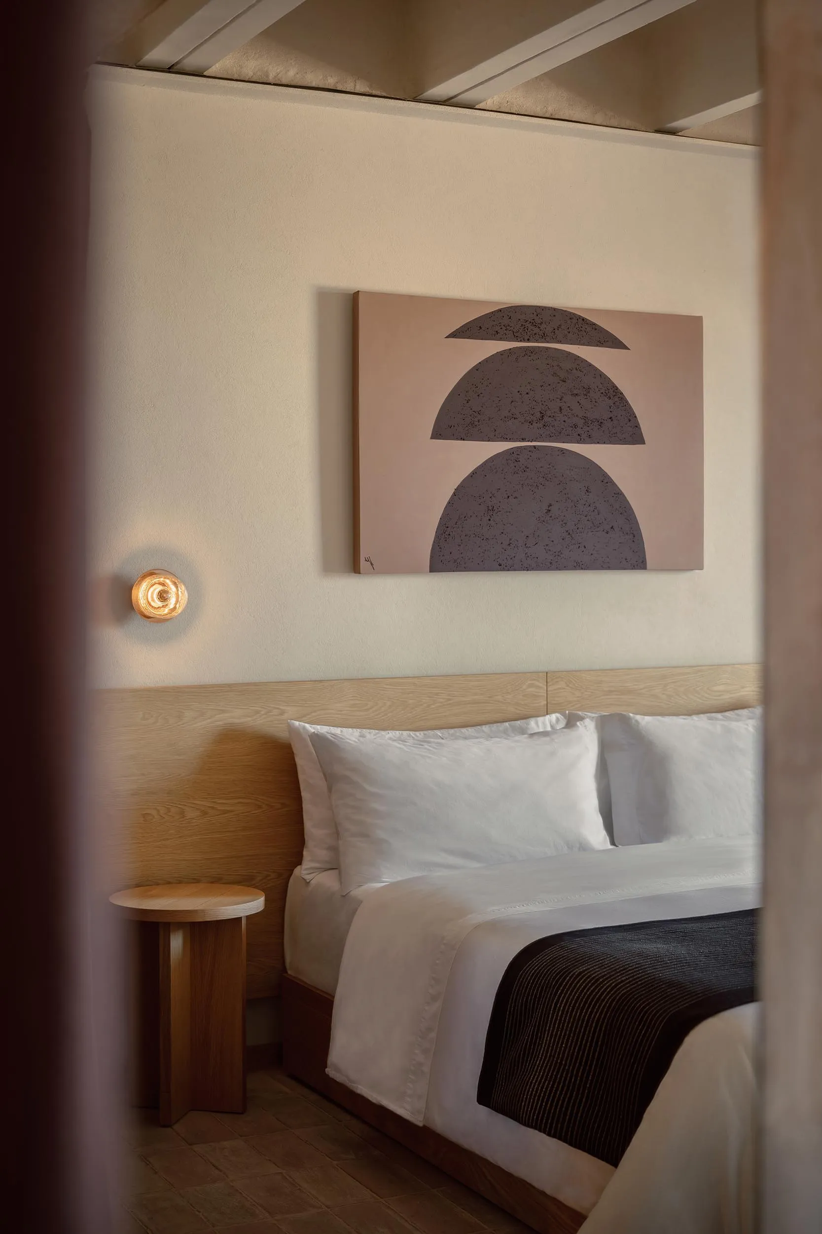A minimalist bedroom with a wooden headboard and white bedding, featuring a wall-mounted light fixture and a large abstract p