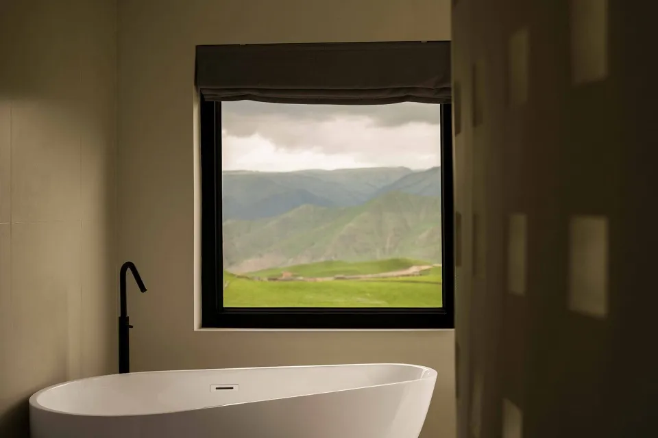 A modern bathroom featuring a white freestanding bathtub with a view of green hills and mountains through a large window with