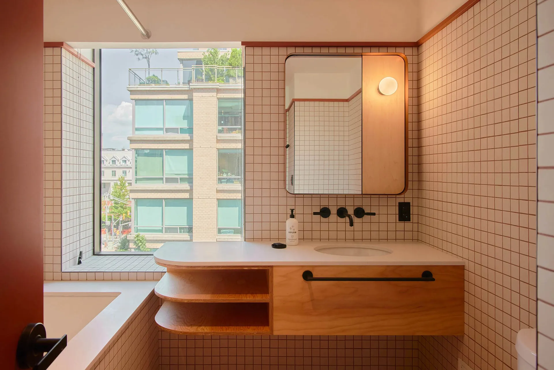 Modern bathroom with white ceramic tiles, a floating wooden vanity, and a large window showcasing urban architecture.