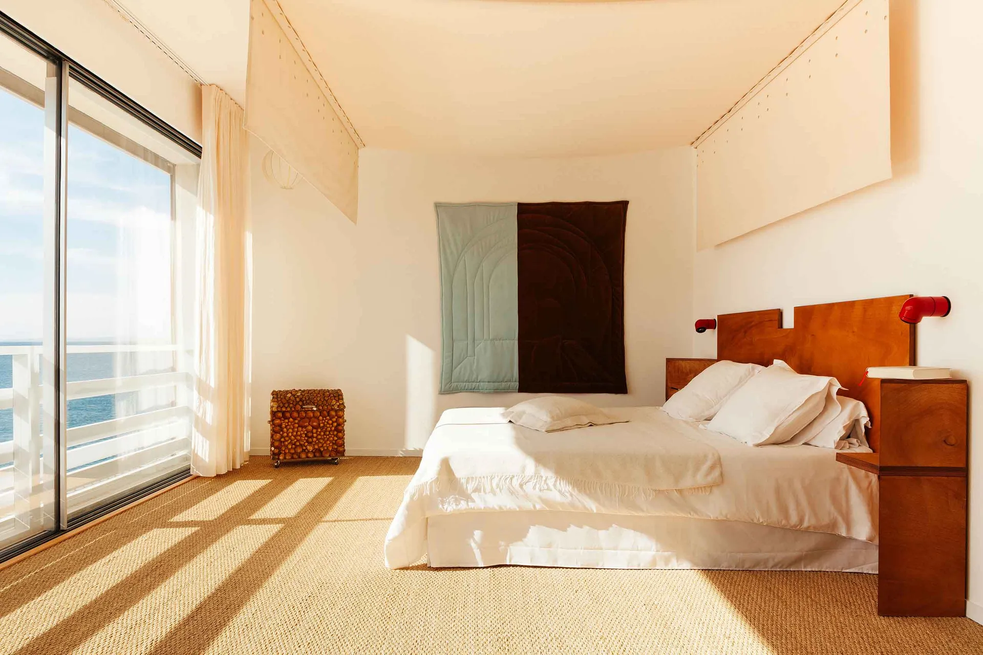 Minimalist bedroom with a bed, wooden headboard, and side tables, featuring a large window with a view of the ocean.