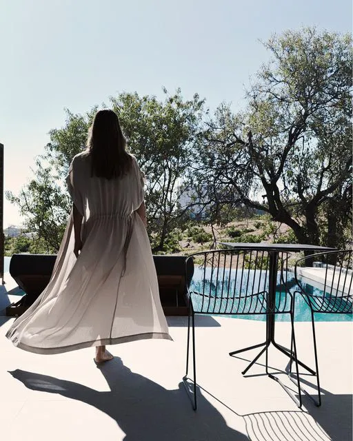 A woman in a long dress walks on a patio with concrete, metal, and glass elements, overlooking a pool and trees.