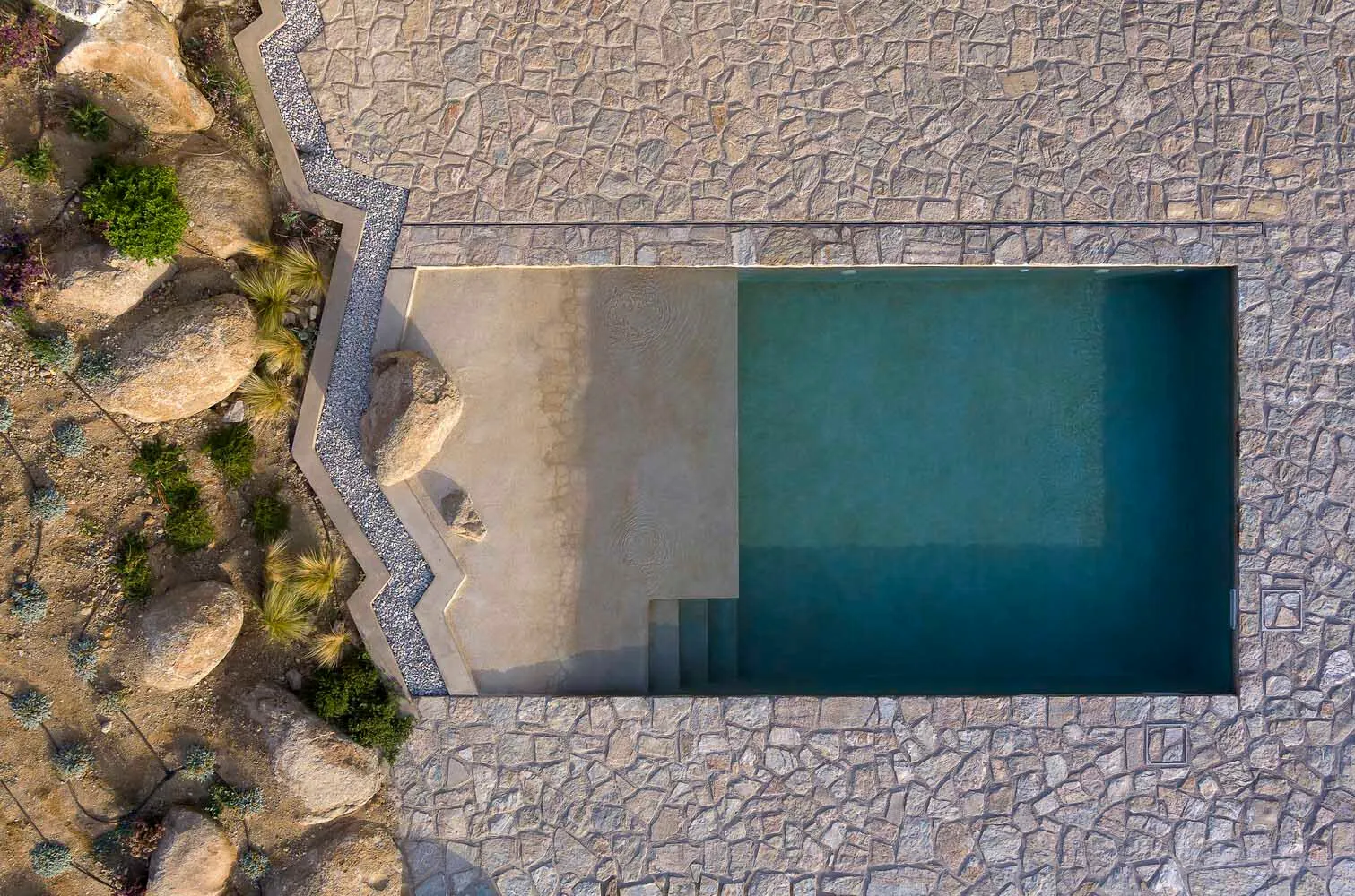 An aerial view of a modern pool with concrete, rock, and gravel elements.