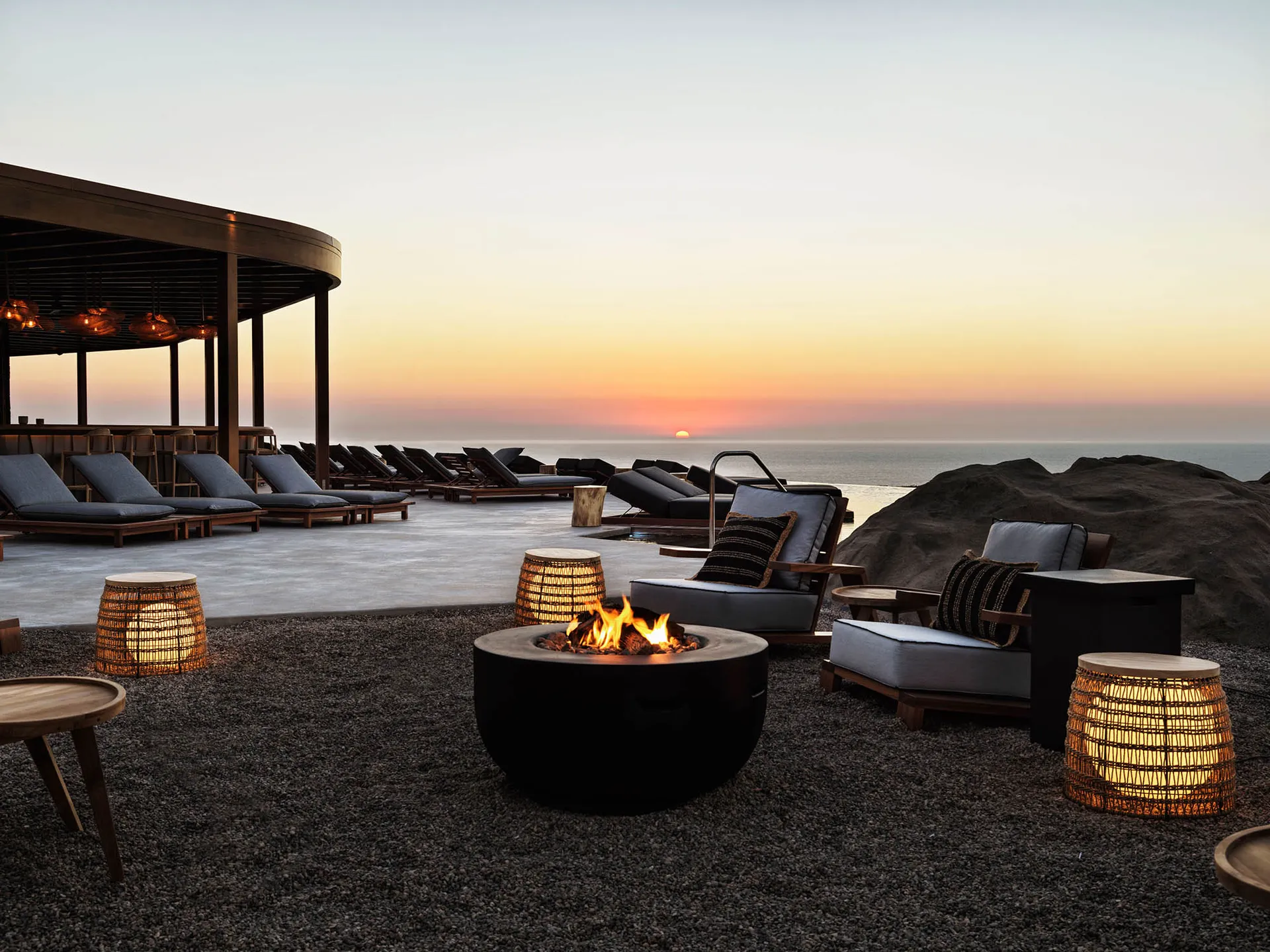 Outdoor lounge with concrete seating, wooden chairs, and wicker stools, overlooking the ocean during sunset.
