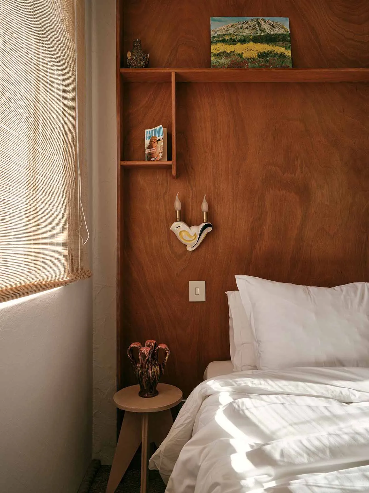 A minimalist bedroom features a bed with white bedding, a wooden headboard, and a wall-mounted sconce. The room is adorned wi