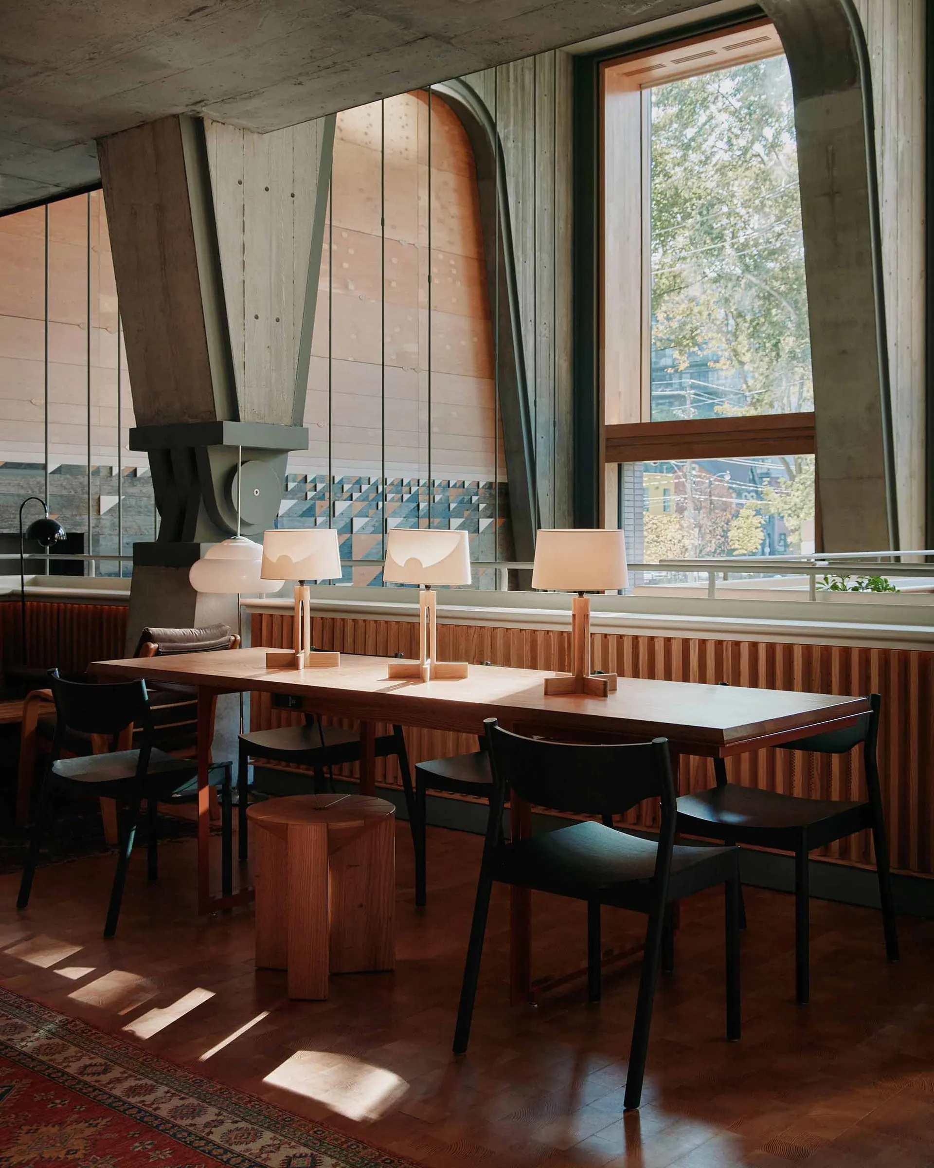 A modern desk space featuring a long wooden table with three table lamps and black chairs, surrounded by large windows with v