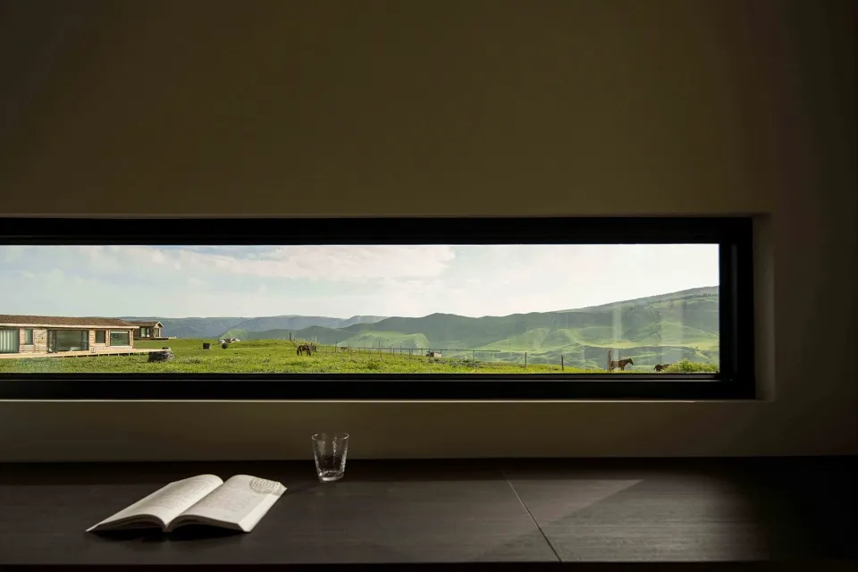 A modern window with a view of green mountains and a house in the background.