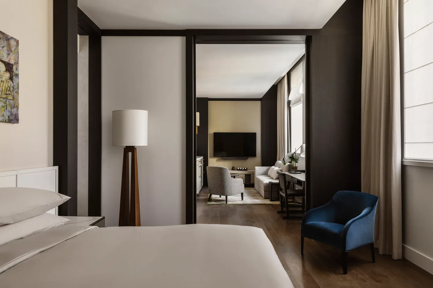 A modern hotel room featuring a minimalist bedroom with a bed, lamp, and a door leading to a living space.