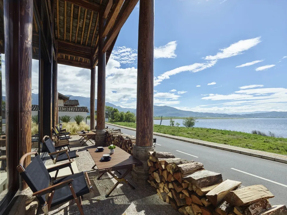 Outdoor terrace of a resort with wooden tables, chairs, and firewood, overlooking a serene lake and mountains under a clear b