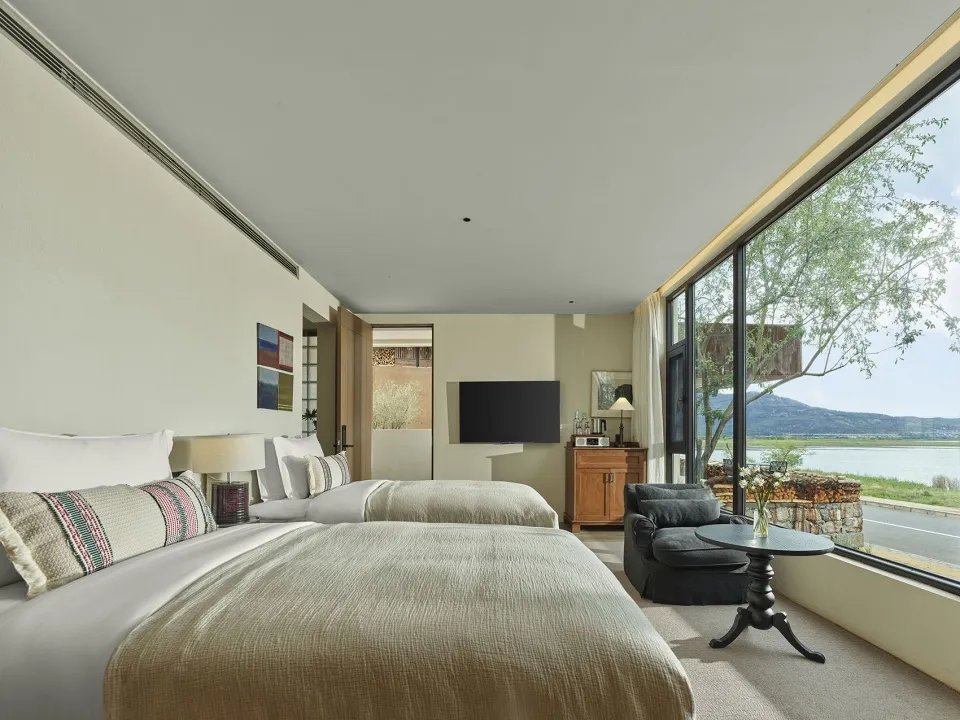 A modern minimalist bedroom with two twin beds, a flat-screen TV, and a view of the water.