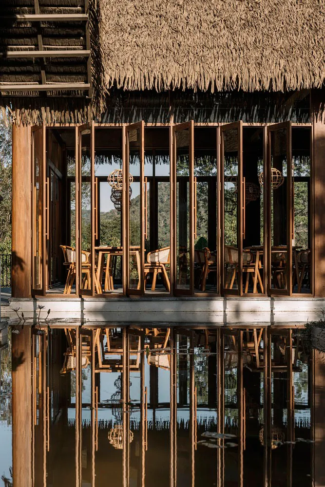 A rustic restaurant with a thatched roof, open folding doors, wooden furniture, and a scenic water reflection.