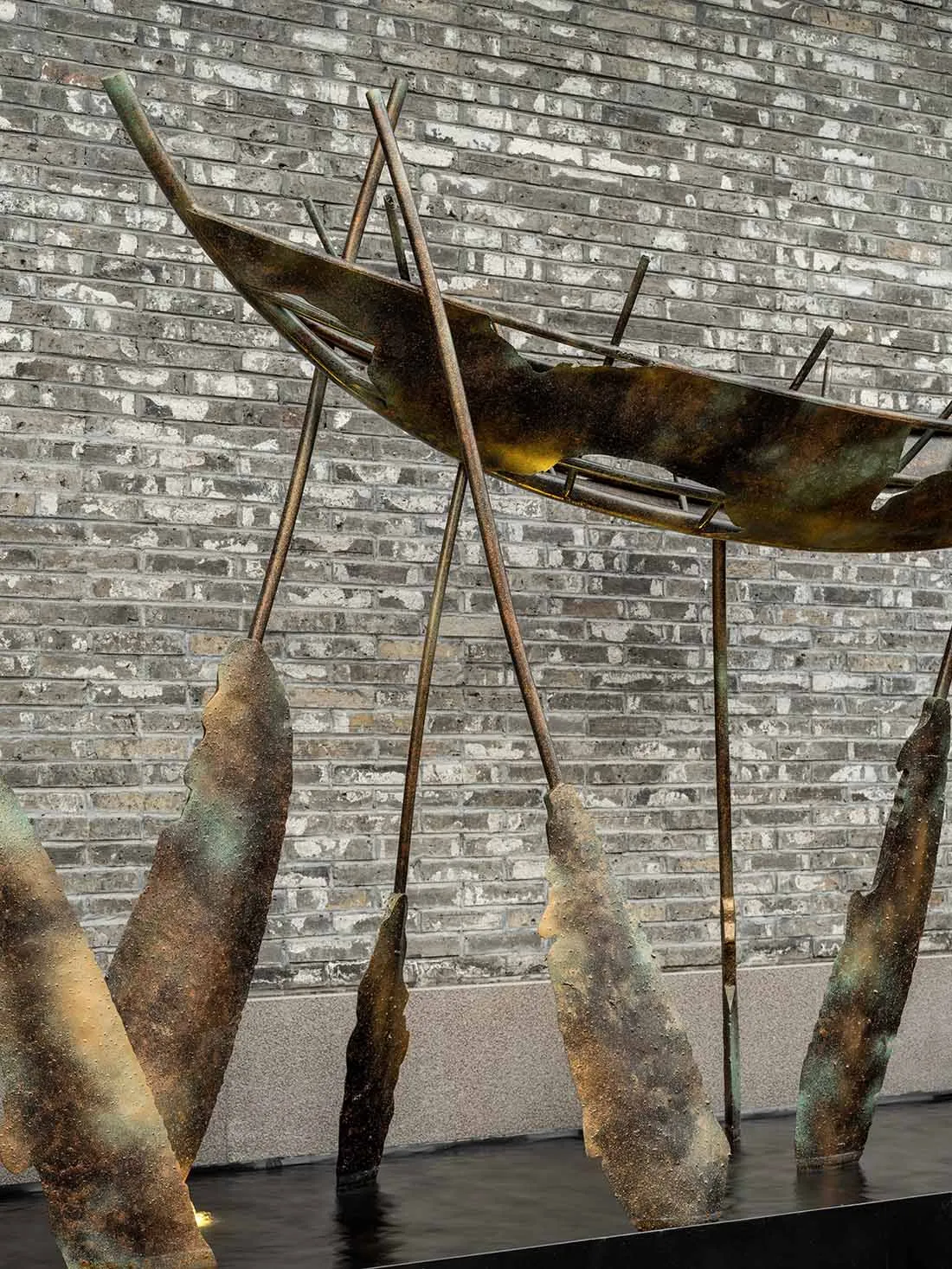A bronze abstract sculpture with crossed metal rods and water elements in front of a brick wall.