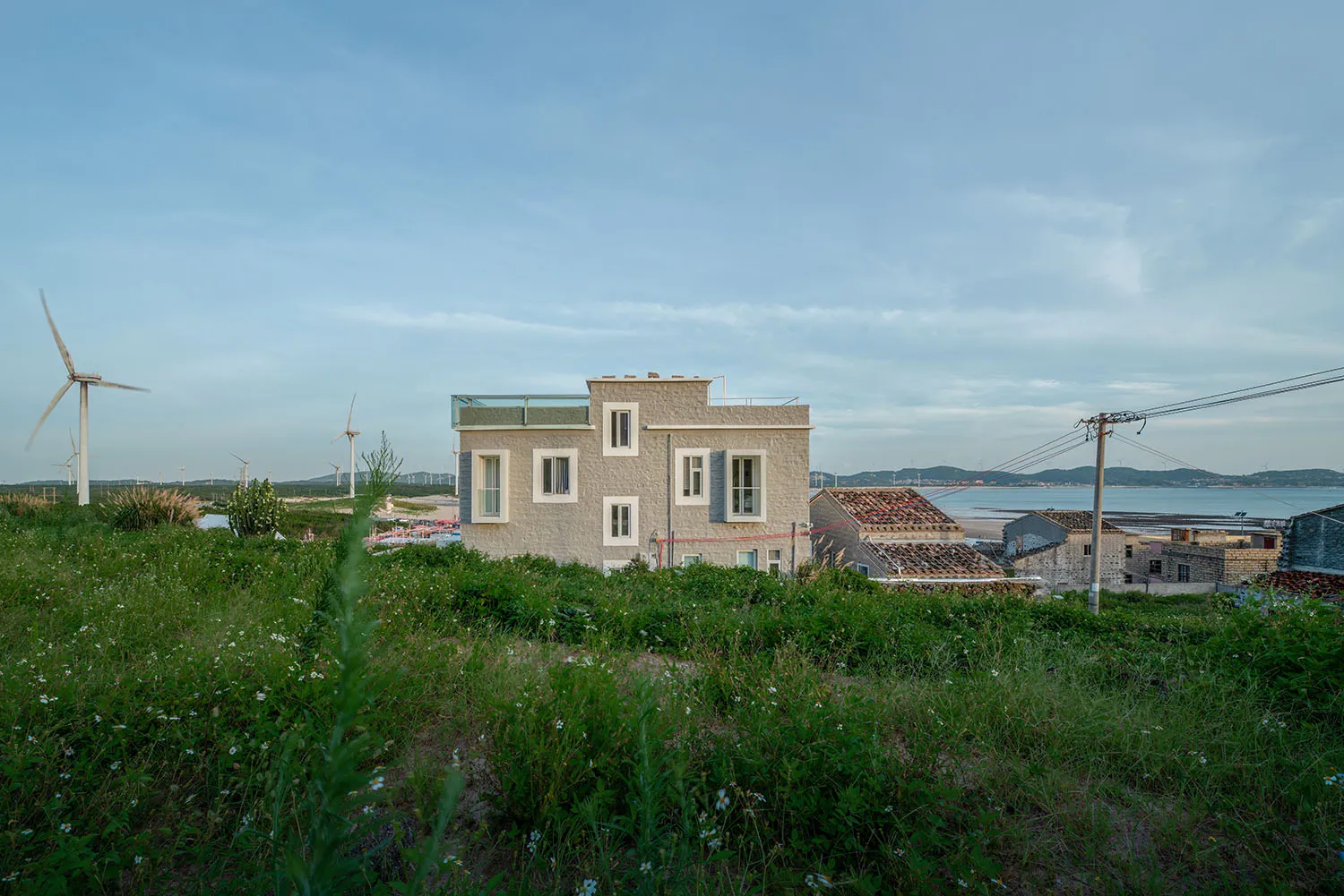 A minimalist-style beach house with concrete walls, surrounded by greenery and wind turbines.