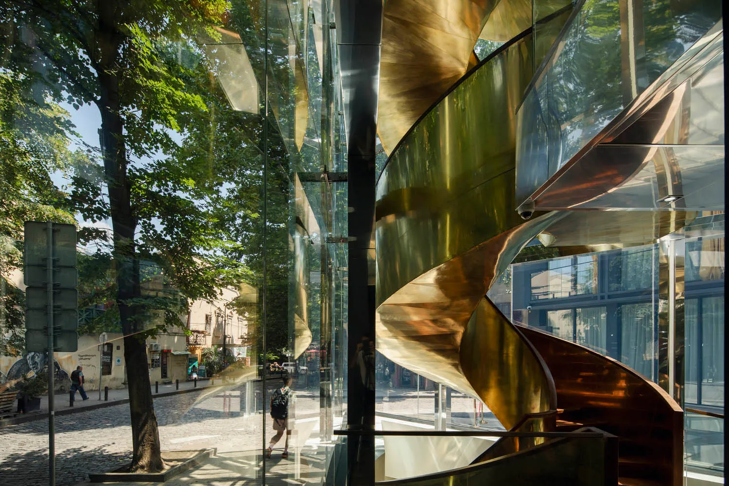 A modern building with spiral glass staircase in a sunny urban setting.