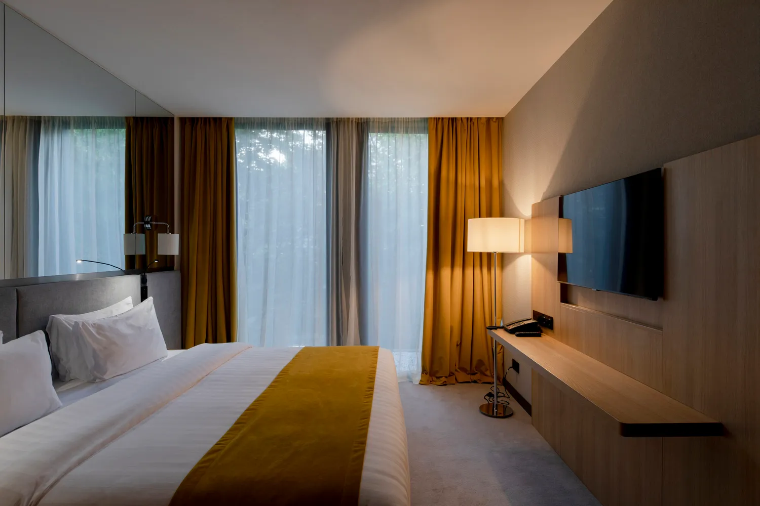 A modern minimalist bedroom with white and gold accents, large windows, and a comfortable bed.