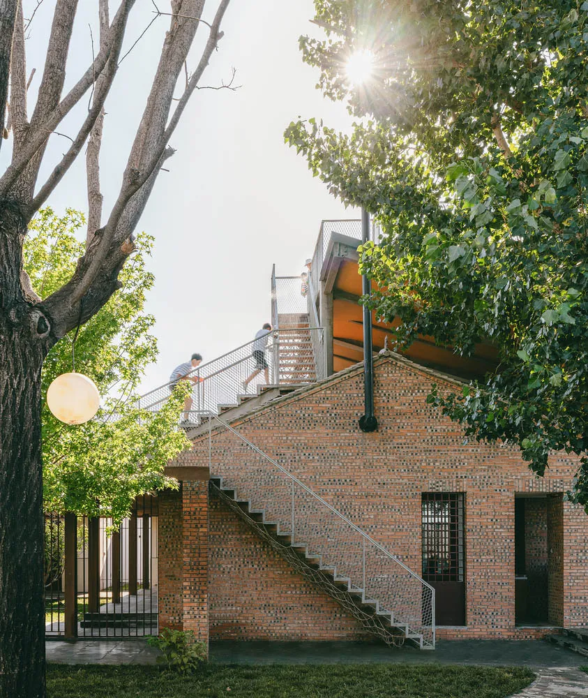 A modern brick house with sunlight shining through trees and a metal staircase leading up to the second floor.