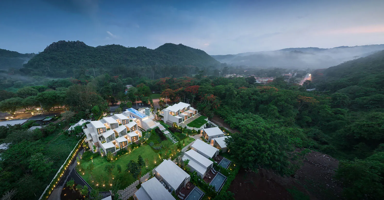 俯瞰一座現代住宅，坐落於綠意盎然的景觀中，背景為山巒。
