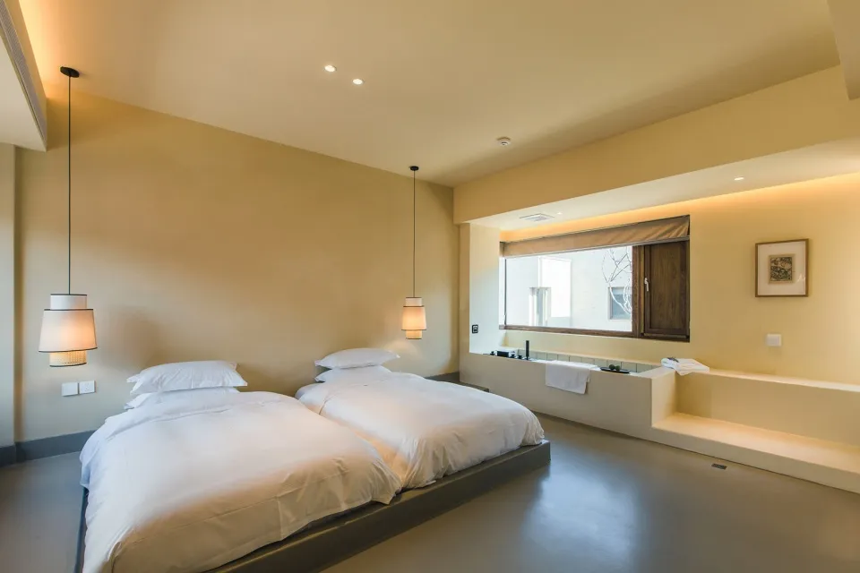 A minimalist bedroom with neutral tones features twin beds, pendant lamps, and a window.