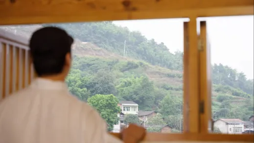 Man standing at window overlooking a mountain landscape with green hills and scattered houses.