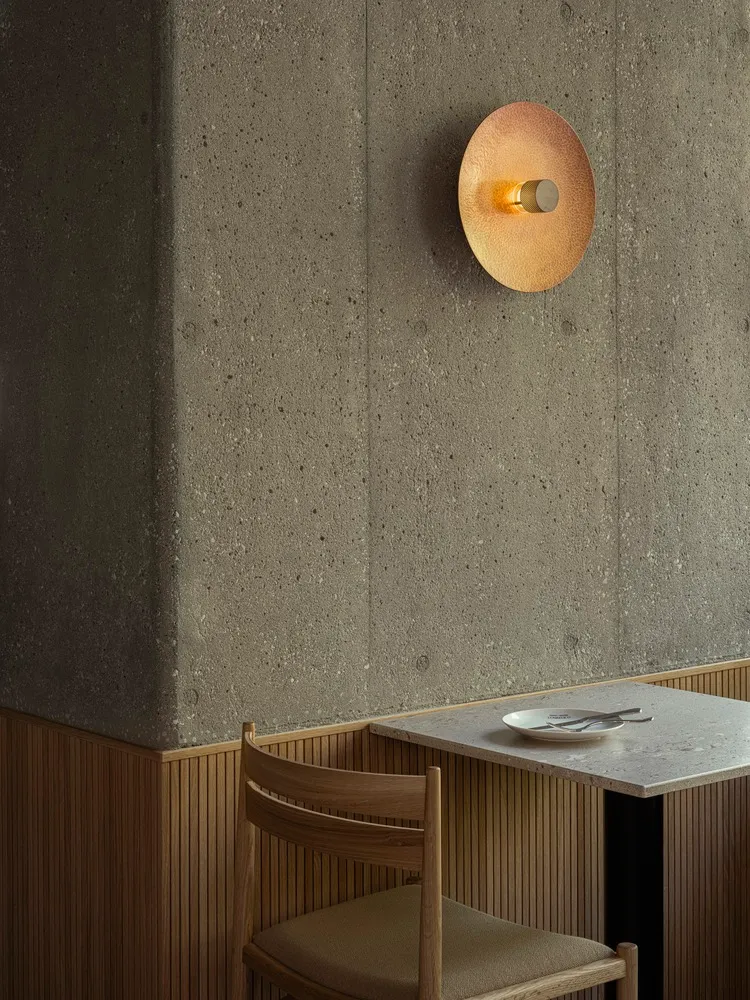 Minimalist restaurant interior featuring a concrete wall, wooden chair, and round gold light fixture.