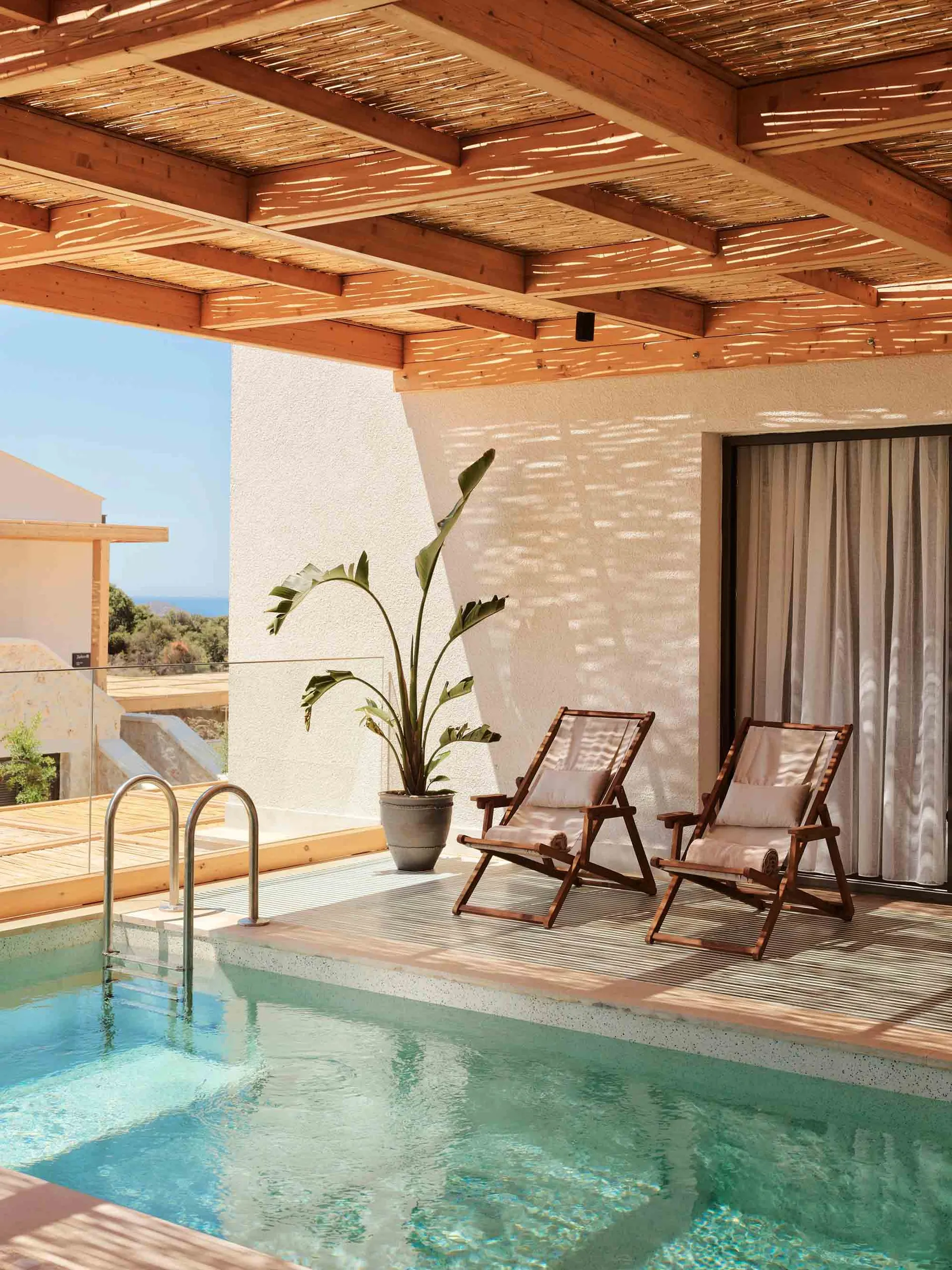 A private living space featuring a swimming pool, wooden deck chairs, and a potted palm plant under a wooden pergola.