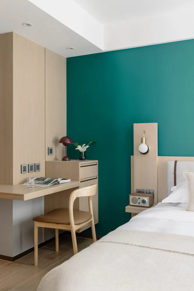 A modern minimalist bedroom featuring a green accent wall, wooden furniture, and a white bed.
