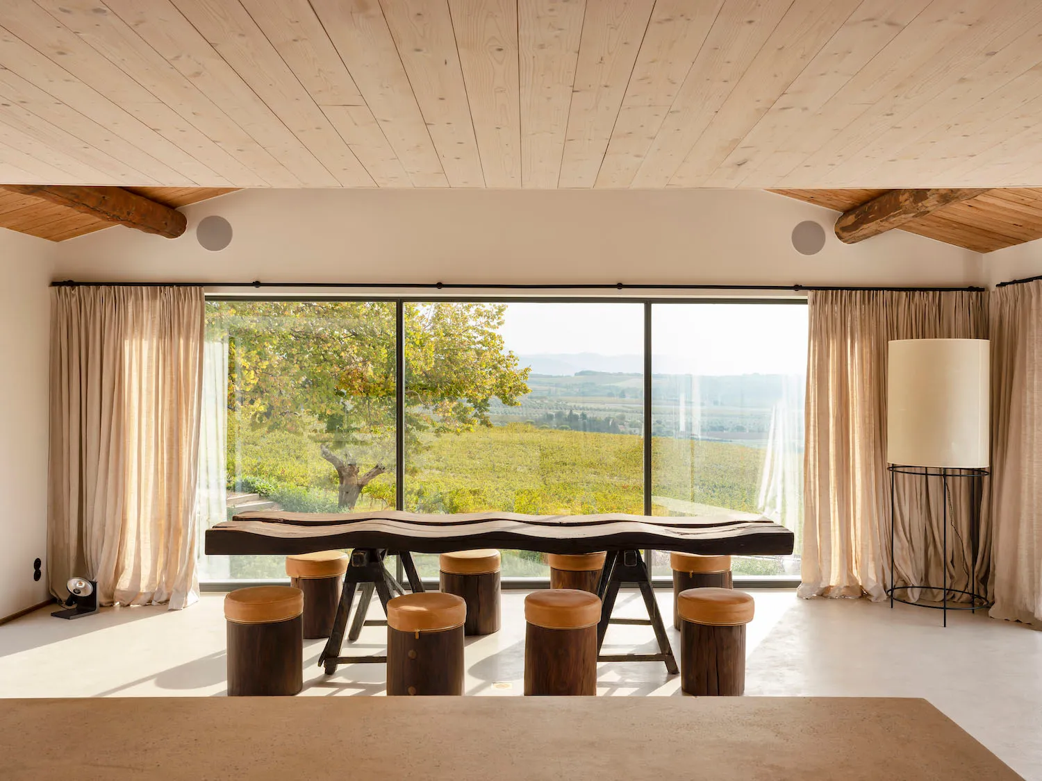 Modern dining room with minimalist design featuring a wooden table and stools, large window overlooking a scenic vineyard.