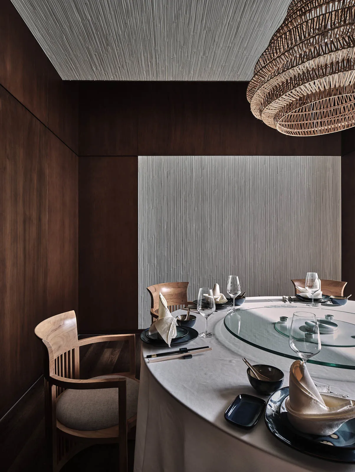 A minimalist restaurant dining room with dark wood walls, glass table, textured fabric chairs, and a woven rattan basket pend