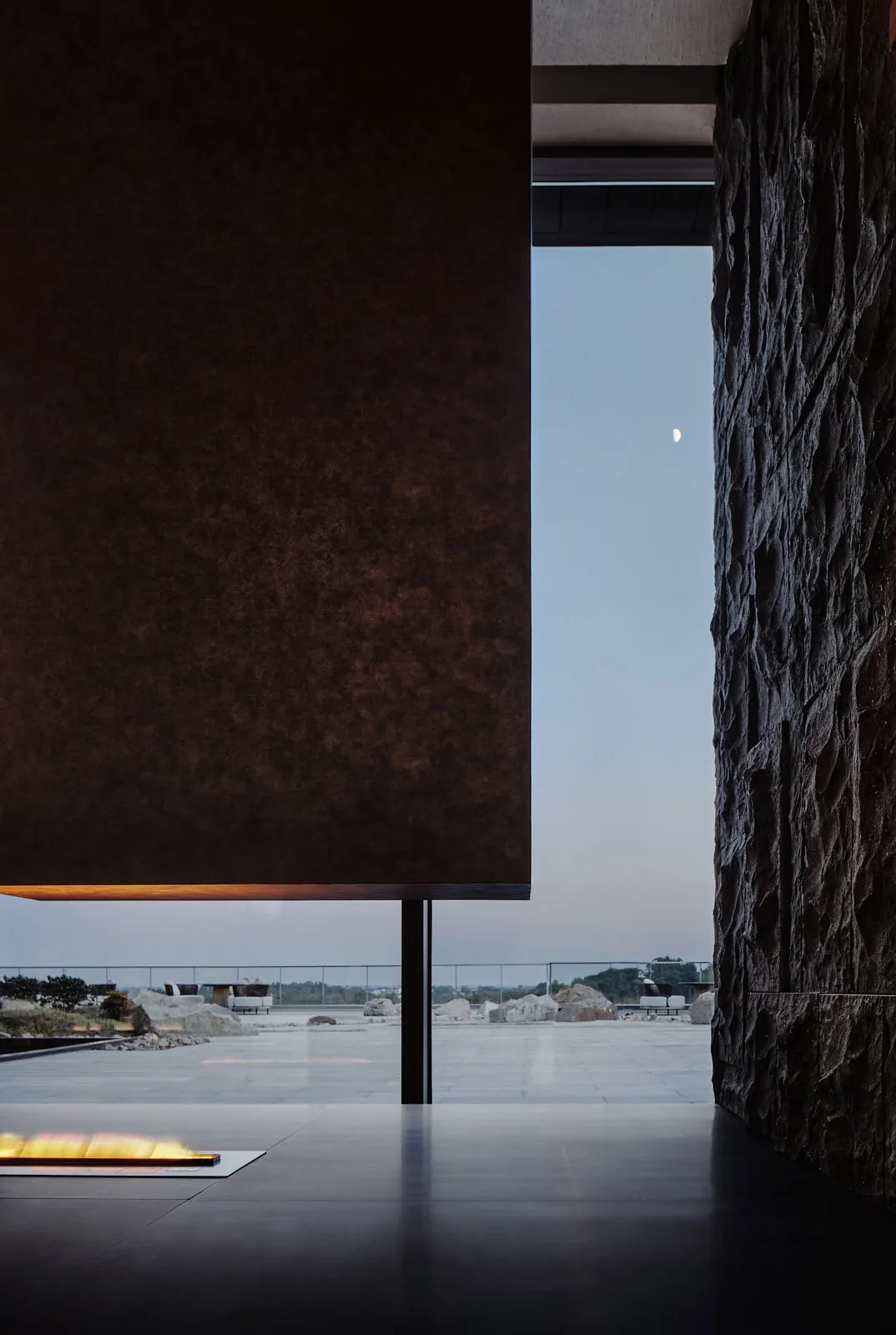 A modern minimalist living room featuring concrete and rock walls, with a minimalist fireplace lit and a view of the outdoor
