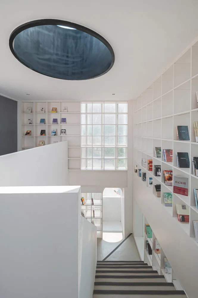 A modern staircase with a bookshelf on a white background, featuring a unique circular skylight.
