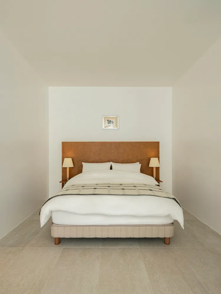 A minimalist bedroom featuring a neutral-toned bed with a simple headboard, side tables with lamps, and a framed picture on t
