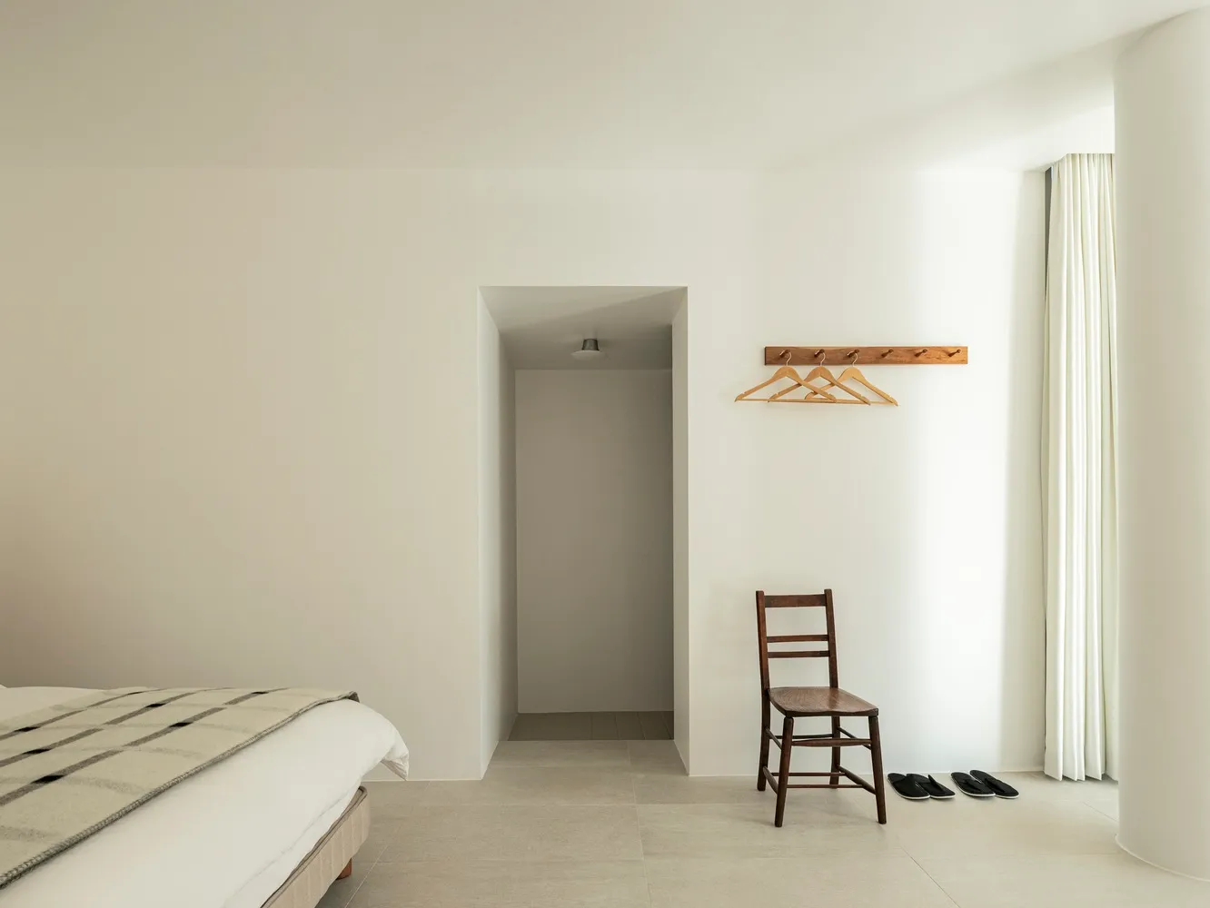 A minimalist bedroom with a white wall, a wooden chair with a pair of black sandals, and a bed with a beige blanket.