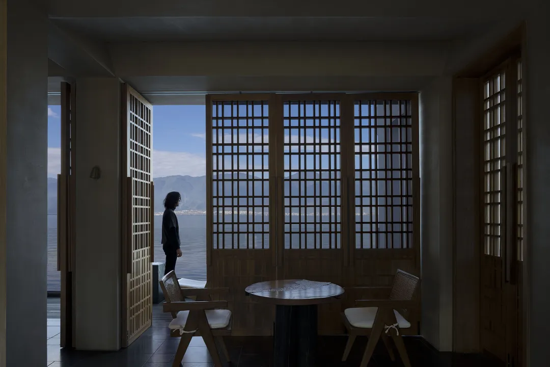 A minimalist interior featuring a view through a window with wooden doors and a table and chairs