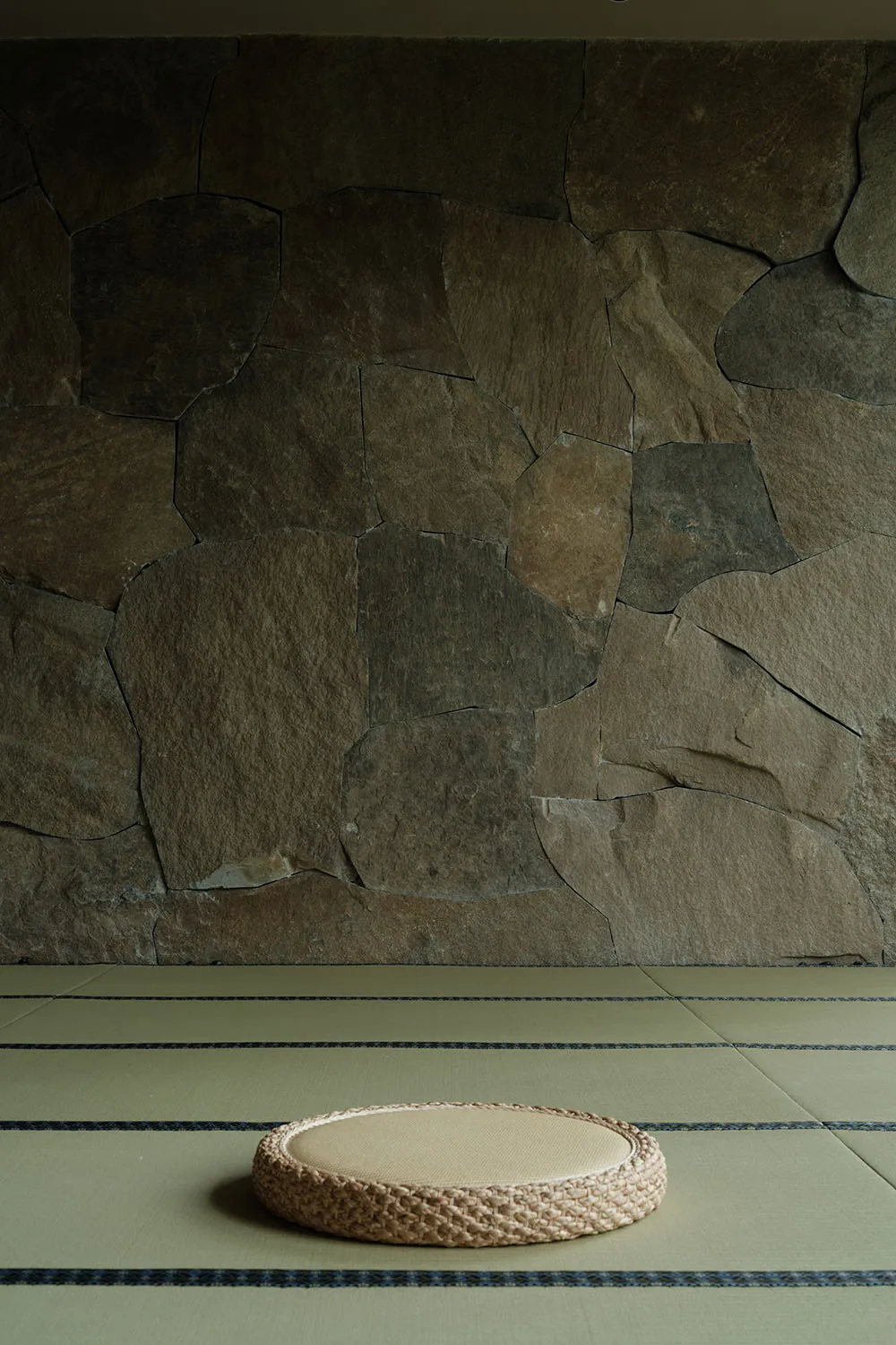 A minimalist floor space featuring a cotton and rattan cushion on tatami matting with a stone wall background.