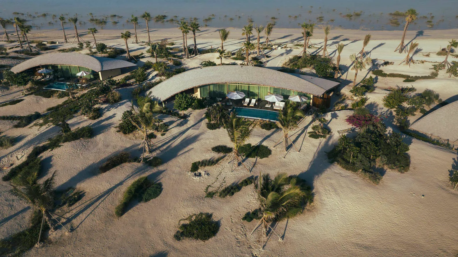 A modern resort with curved architecture, a pool, and palm trees in a sandy environment.
