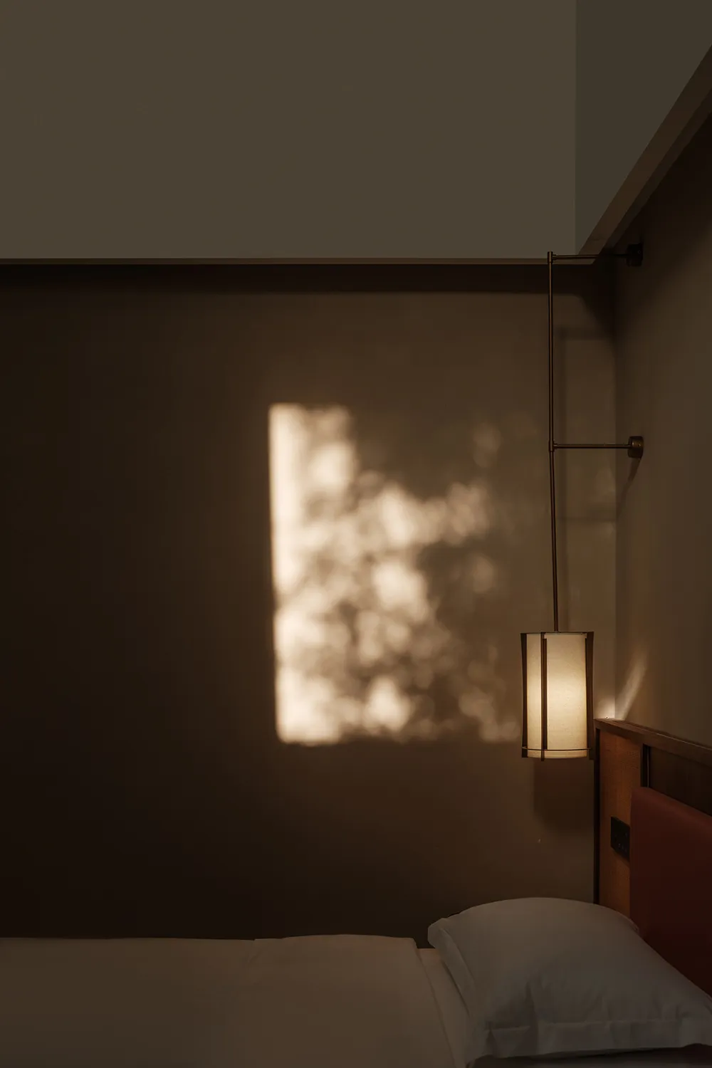 A minimalist bedroom with a grey wall, a bed with white bedding, and a hanging light fixture casting a warm glow on the wall.