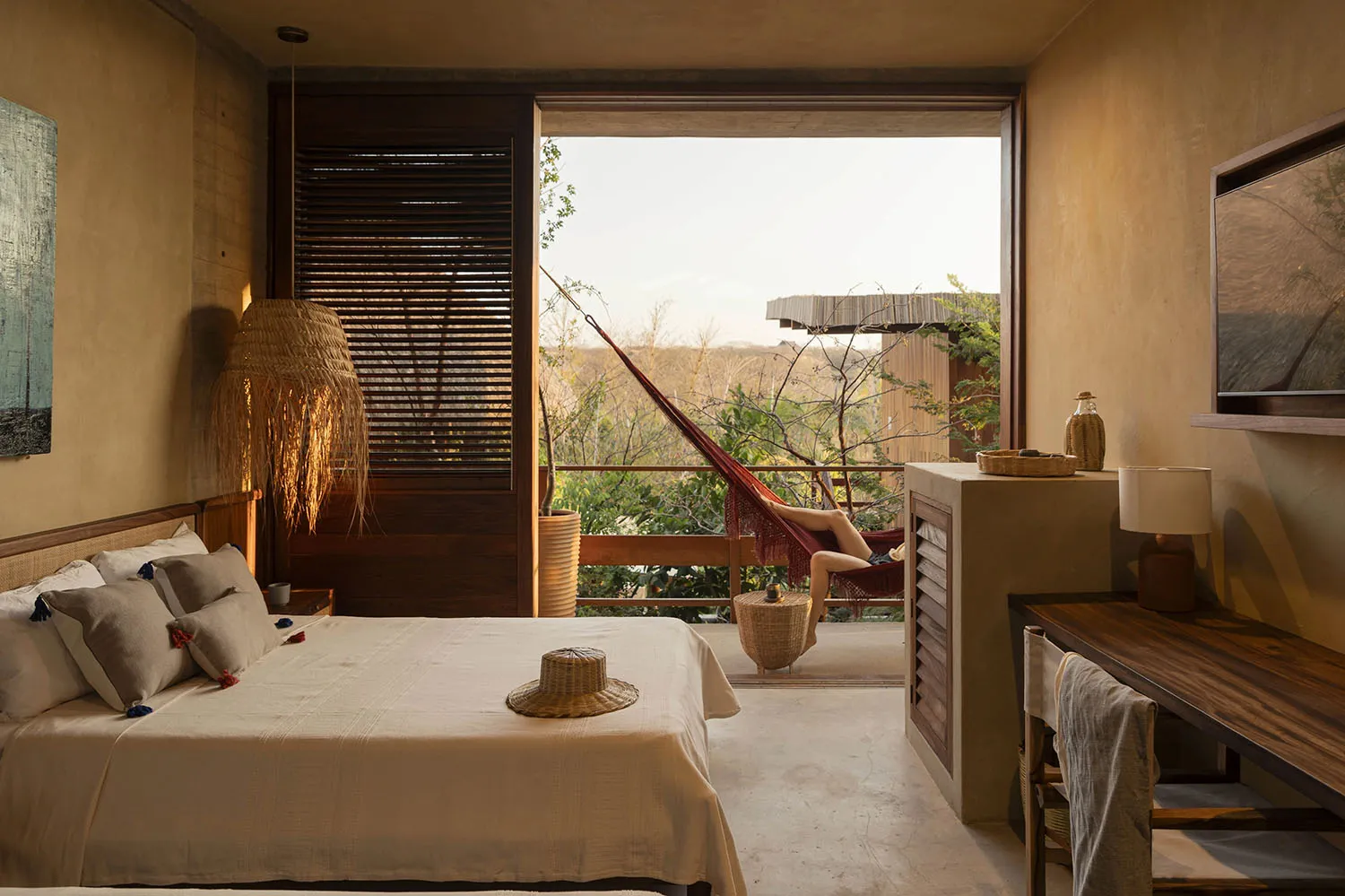 A minimalist bedroom with a bed, bedside table, and a hammock on the balcony overlooking trees.