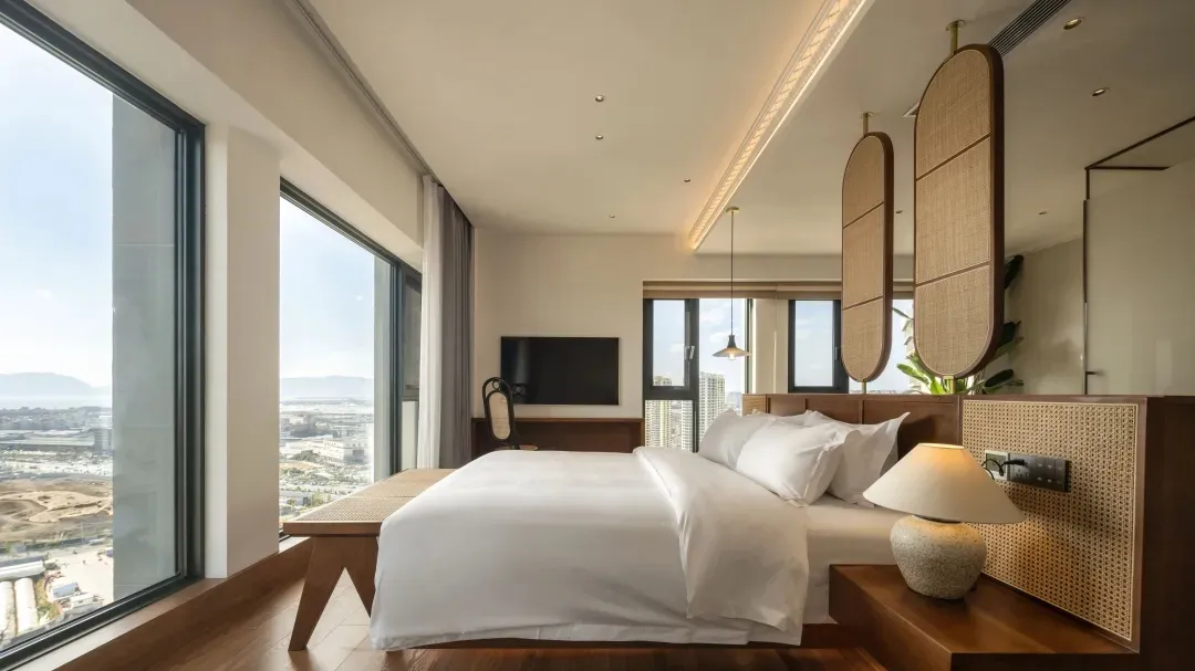 A modern bedroom with a white bed, wooden headboard, and large window overlooking a cityscape.