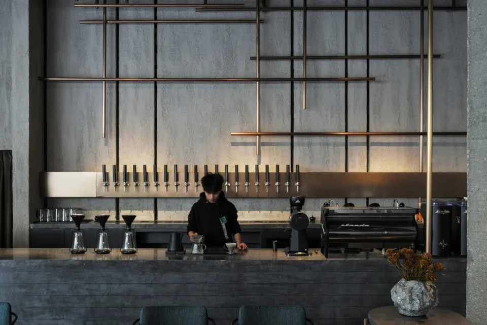A minimalist coffee bar featuring a concrete counter with a person serving coffee, industrial-style lighting fixtures hanging
