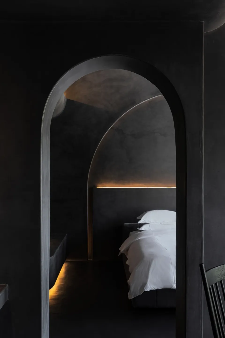 A minimalist bedroom with a concrete headboard and floor, featuring a single white bed and soft lighting.