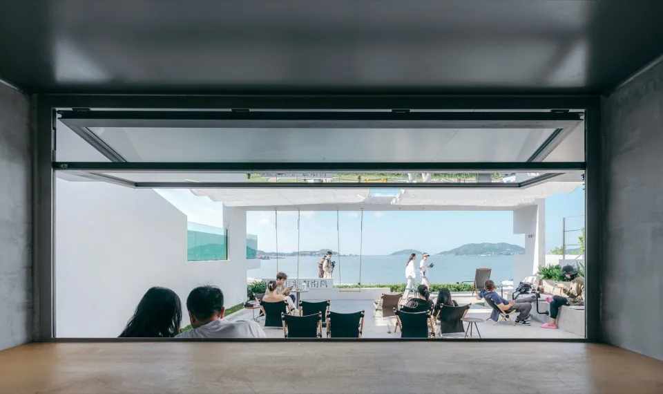 People sitting near a large glass window overlooking the ocean and mountains.